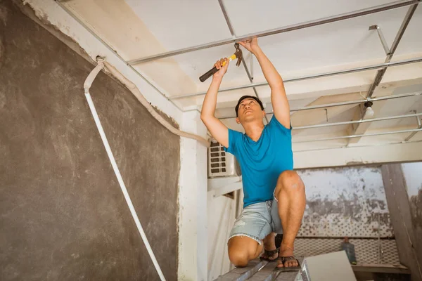 young Asian man hammers in nails while repairing a ceiling frame during roof renovations