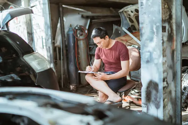young male mechanic sitting while writing a list of car repairs at a repair shop