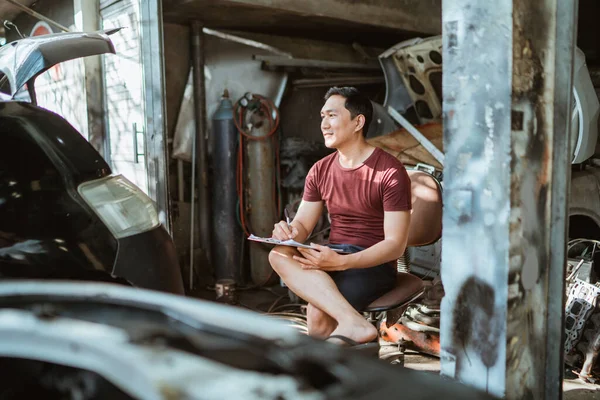 young male mechanic sits recalling while writing a list of car repairs at a repair shop