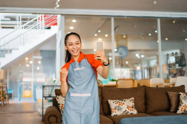 smiling shop assistant woman in apron selfi with cell phone standing in front of furniture store