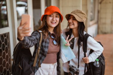 Arka planda evi olan cep telefonu kamerasıyla selfie çeken iki güzel turistin portresi.