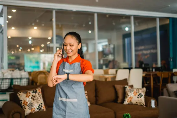 female shop assistant in apron calling on cell phone when seeing watches standing in front of furniture store