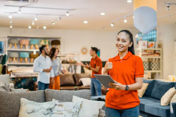 female store clerk with thumbs up holding a tablet standing against the background of shoppers and shopkeeper in furniture store