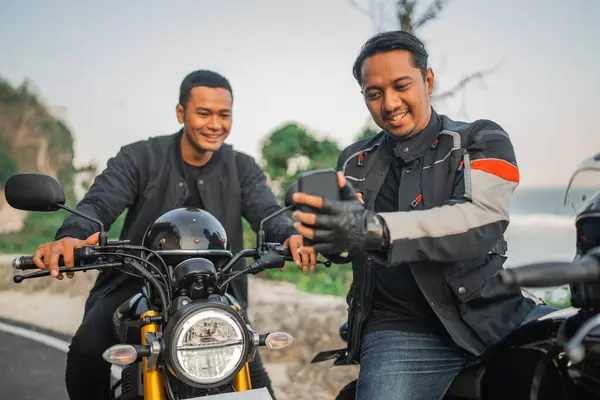 アジアのモーターサイクリストは 携帯電話を持って 彼の友人と一緒にそれを見ています — ストック写真