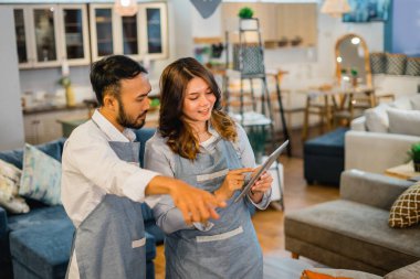 Önlüklü kadın ve erkek tezgahtarlar mobilya mağazasında eşyaları kontrol ederken birlikte tablet kullanıyorlar.