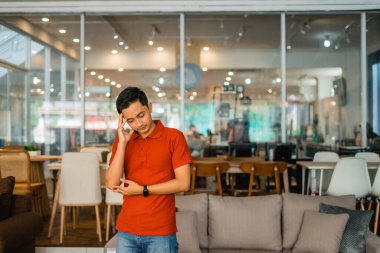 Kırmızı elbiseli erkek tezgahtar, mobilya mağazasında sersemlerken başını tutuyor.