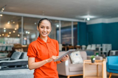 Elinde tablet olan gülümseyen genç bir kadın mobilya mağazasının önünde dikiliyor.