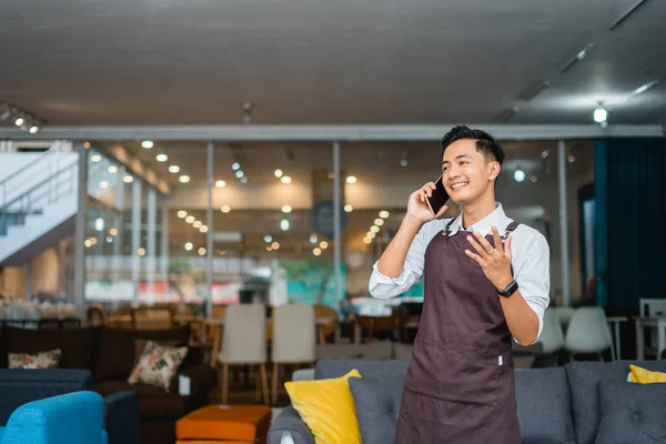 male shop assistant in apron making a cell phone call standing in front of furniture department store