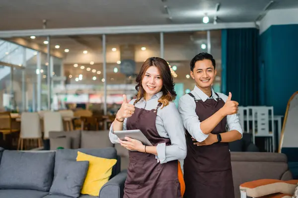 Asian male and female waiter in apron smiling with thumbs up while holding a tablet in front of a furniture store