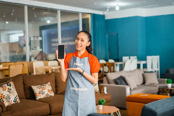 female shop assistant in apron with hand gesture presenting cell phone in front of furniture store