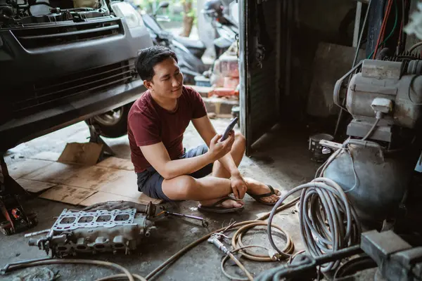 Young mechanic using a cell phone while sitting on the floor resting after repairing an engine in a garage