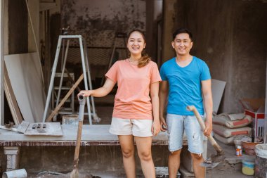 Mutlu genç çift yeni bir ev inşa ediyor. Evin yanında kürek ve fincanla duran karı koca ve hammaddeler.