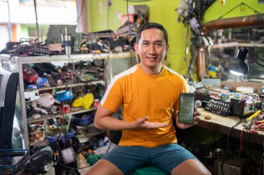 Asyalı elektronik tamircisi, bir elektronik tamir dükkanında cep telefonu ekranını gösterirken gülümsüyor.