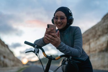 tesettüre binen bisikletli, telefon ve kulaklık kullanan, müzik ya da şarkı dinleyen