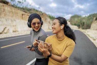 Spor giyimli iki kadın gülüyor, telefon kullanıyor, yolda koşuyor.