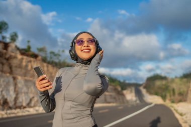 Gülümseyen tesettürlü kadın spor kıyafetleri giyiyor. Müzik dinlemek için telefon ve kulaklık kullanıyor.