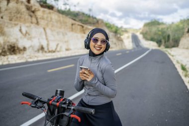 Kadın motorcu tepede bisiklet sürerken kulaklık ve telefon kullanıyor.