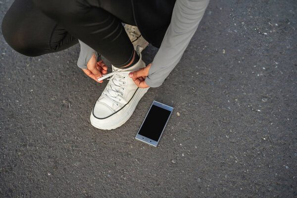 обрезанный женщина в положении приседания завязывая шнурки с пустым экраном телефона