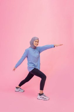 Canlı spor kıyafetleri içinde bir kadın zarif bir şekilde pembe bir arka plana yaslanıyor.