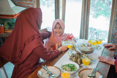 Küçük tesettürlü kız anne ve ailesiyle yemek masasında iftar yaparken mutlu bir şekilde gülümsüyor.