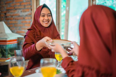 Sıcak bir ortamda, renkli tesettürlü iki kadın bir yemeği paylaşıyor, birlikte vakit geçiriyorlar.