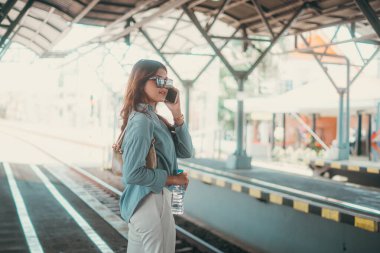 Güneş gözlüklü şık bir kadın meşgul, hareketli bir tren istasyonunda telefonla konuşuyor.