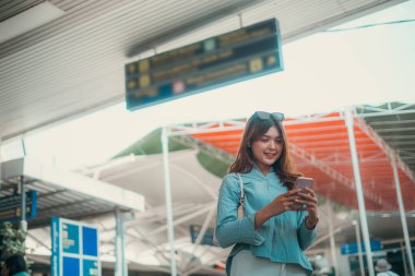 A joyful traveler happily enjoying her time at the bustling airport while using her phone clipart