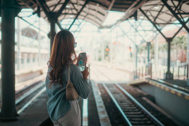 Yoğun bir tren istasyonunda duran bir kadın, akıllı telefonuna odaklanmış, trenini bekliyor.