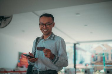 Evde gülümseyen genç bir adam elinde kredi kartıyla telefonunu kontrol ediyor.