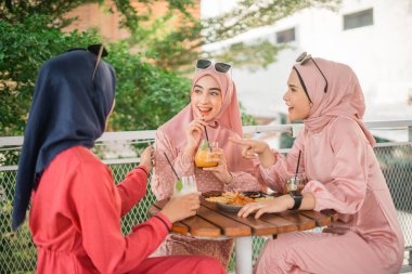 Renkli tesettürlü üç arkadaş güneşli bir kafede içkinin ve kahkahanın tadını çıkarıp, neşeyi paylaşıyorlar.