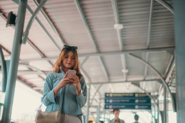 Genç bir kadın, kalabalık bir şehir taşıma istasyonunda insanlarla çevrili akıllı telefonuna odaklanır.