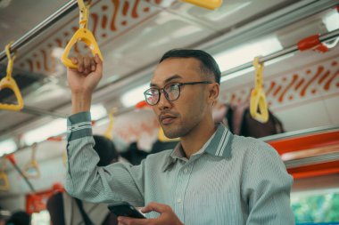 Dikkatini telefon ekranına odaklayan bir adam hareket halindeki bir trenin korkuluğunu sıkıca tutuyor.