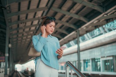 Bir kadın hareketli bir tren istasyonunda duruyor, dikkatle telefonuna bakıyor ve trenlerinin gelmesini bekliyor.
