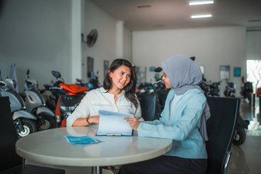 Genç kadın ve peçeli satıcı galeriden yeni bir motosiklet almadan önce onay mektubunu okurken sohbet ediyorlar.