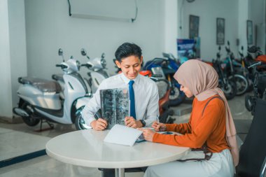 Motosikletli satıcı galeride broşür ve fiyat listesine bakarken peçeli bir kadın müşteriye hizmet veriyor.