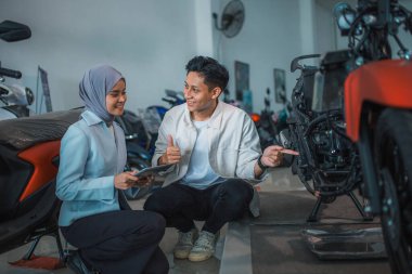 Heyecanlı adam ve peçeli satıcı elinde tabletle bir galeride motosikletin altını kontrol ediyor.