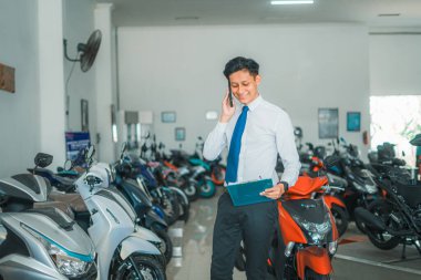 Motosiklet galerisinde erkek satış müdürü arıyor ve elinde motosiklet fiyat listesi var.