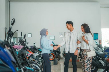 Genç çift, galeriden yeni bir motosiklet almayı kabul ederken peçeli bir satıcıyla tokalaşıyor.