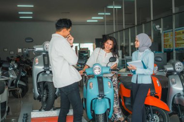 Yeni motosiklet deneyen genç bir kadın ve erkek arkadaşı galeride tesettürlü satıcı tarafından servis ediliyor.