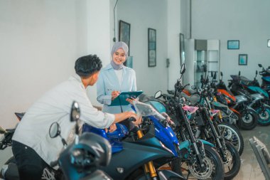 Tezgahtar kadın, galeride motosiklete bakan bir erkek müşteriyle tesettür takıyor.