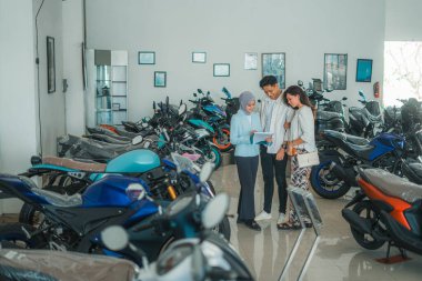 Bir çift, peçeli bir satıcıyla galeride motosiklet fiyatları hakkında sohbet ediyor.