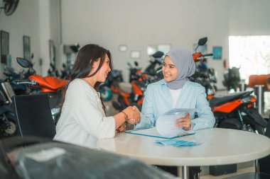 Bir kadın ve peçeli bir bayan tezgahtar, galeride motosiklet satışı anlaşmasında el sıkışıyor.