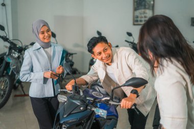 Asyalı bir adam, karısı ve satış elemanı ile motosiklet dükkanında yeni bir motosikletin gidonunu tutuyor.