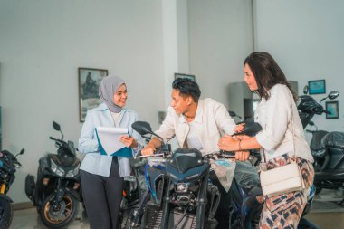 Erkek ve kadın çift yeni bir motosiklet deniyorlar ve tezgahtarlar galeride sohbet ediyorlar.