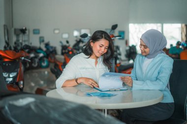 Müşteri peçeli bir bayan tezgahtarla oturuyor ve motosiklet galerisinde sohbet ediyor.