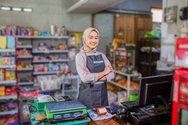 Neşeli ve arkadaş canlısı bir dükkan sahibi hareketli bakkal ortamında kendinden emin duruyor.