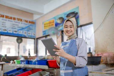 Hareketli ve hareketli bir pazar ortamında önlük takan gülümseyen bir kadın tableti sıkıca tutuyor.