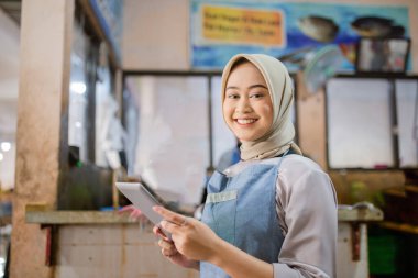 Neşeli bir satıcı canlı ve hareketli bir pazar ortamında dururken tablet kullanıyor.