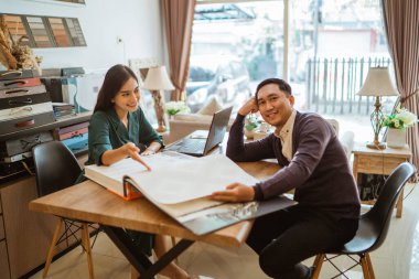 Erkek müşteri kameraya bakarken kadın dükkan sahibi de pencere tedavileri katalog kitabını işaret ediyor.