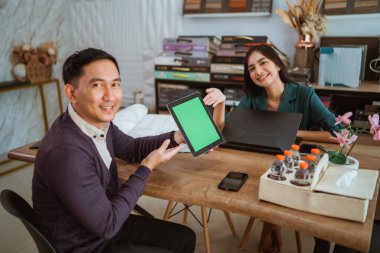 Asyalı kadın esnaf gülümserken, Asyalı iç mimar tarafından tutulan yeşil ekran dijital tableti işaret ediyor.